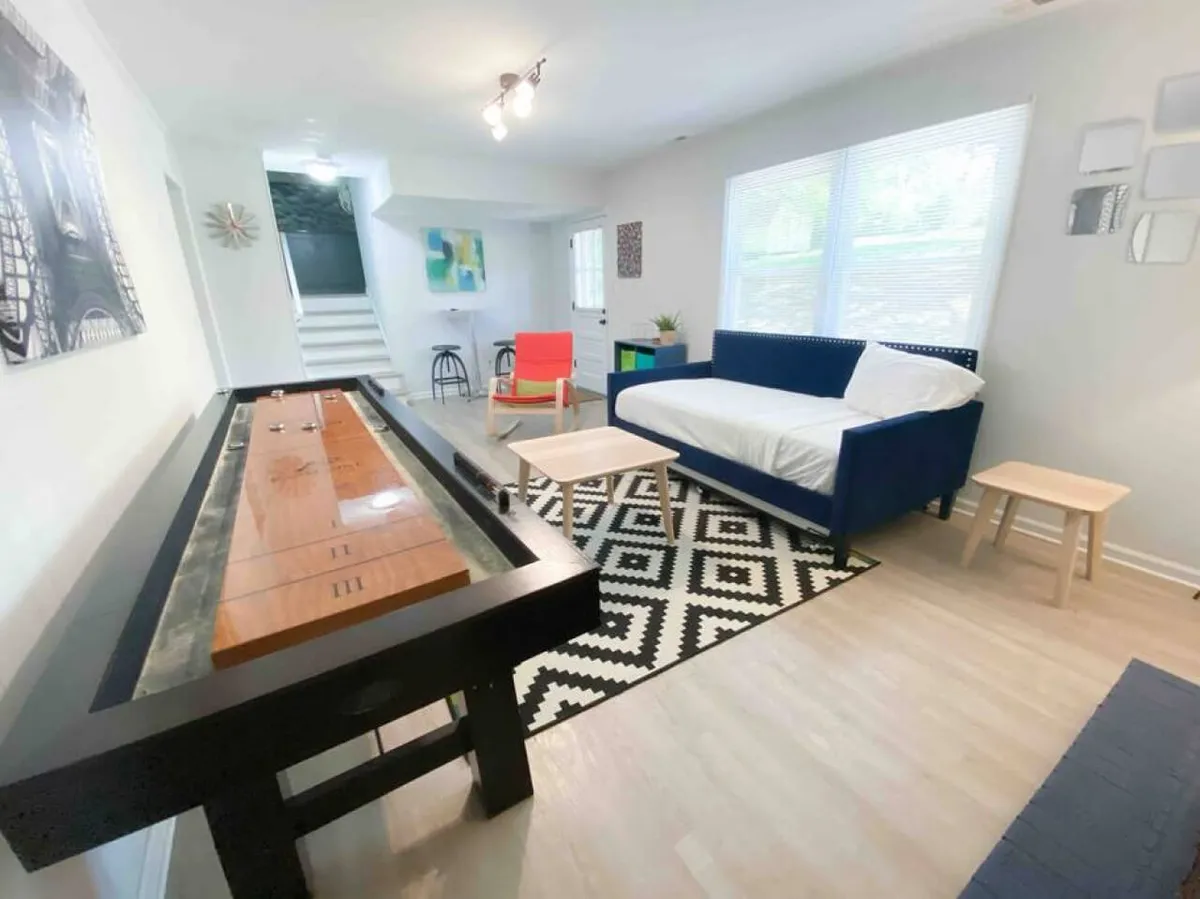 This image centers on a shuffleboard table set against a cozy backdrop with a blue sofa, area rug, and lively wall art. The space encourages playful competition and relaxed lounging, inviting friends to socialize and enjoy shared activities.