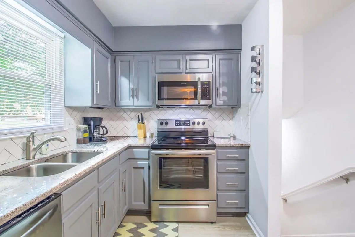 This image displays a stylish kitchen featuring stainless steel appliances, a striking herringbone backsplash, granite countertops, and abundant natural light filtering in through a window. The setup hints at both culinary creativity and communal gatherings.