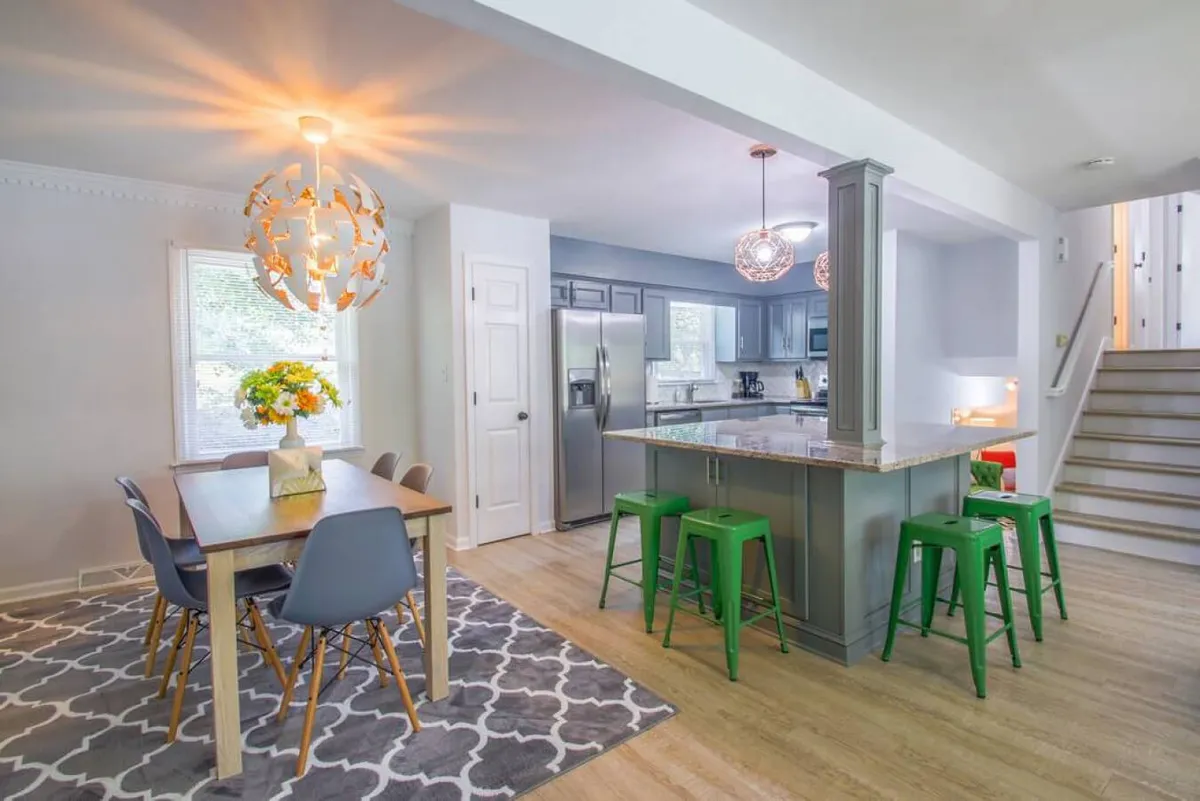 A welcoming dining area is shown here, complete with a dining table adorned with flowers, a kitchen island, unique light fixtures, and colorful bar stools. The space is designed for both intimate family meals and lively gatherings.