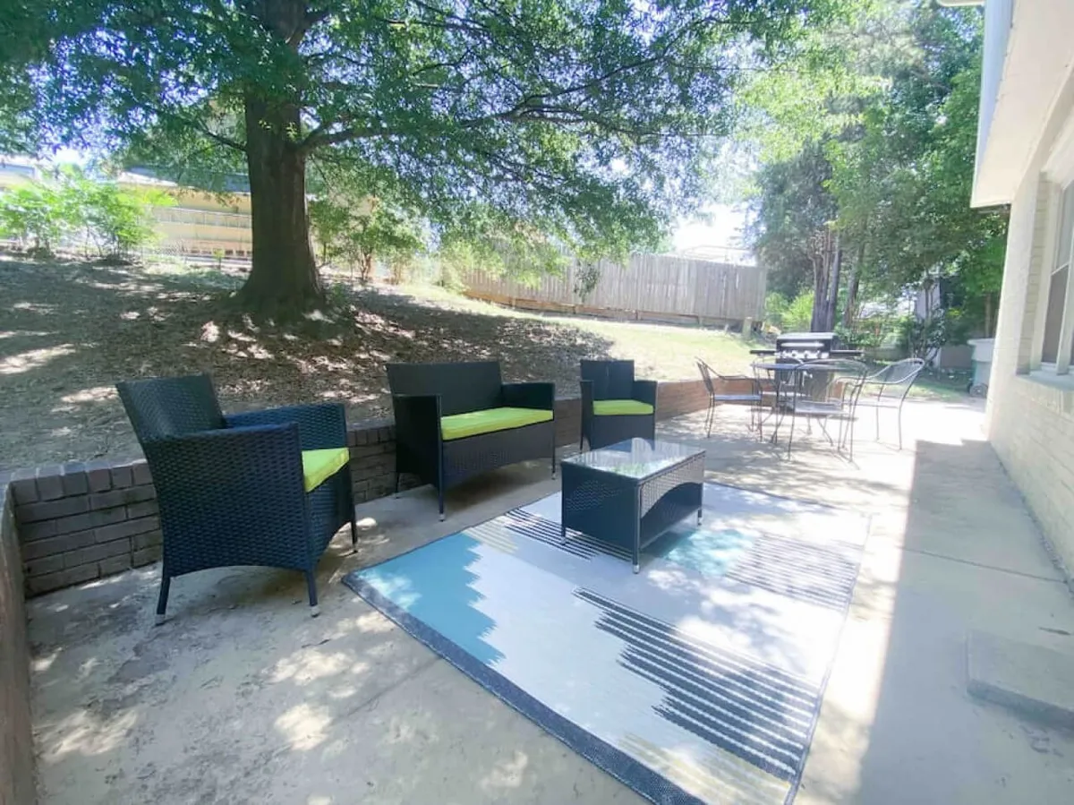 This image features a comfortable outdoor seating area with green cushions, a decorative rug, and lush surrounding greenery. The space invites relaxation and socialization and is bathed in natural light, highlighting its welcoming atmosphere. The setting is ideal for quiet moments with a book or lively conversations, encapsulating the spirit of al fresco living.