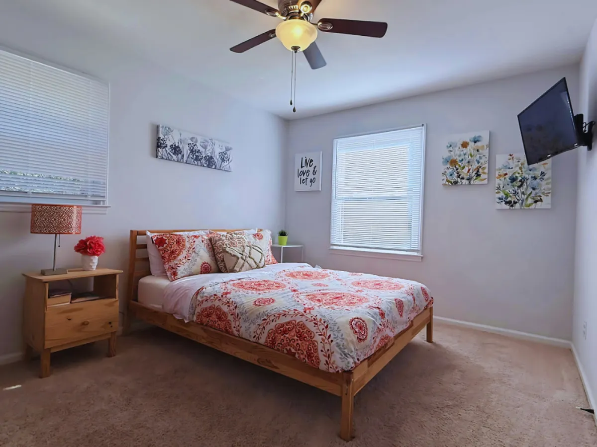 A cozy bedroom featuring a bed with colorful patterned bedding, decorative wall art above the headboard, a nightstand with a lamp and a vase of fresh flowers. The room is bathed in natural light, creating an inviting and restful atmosphere.