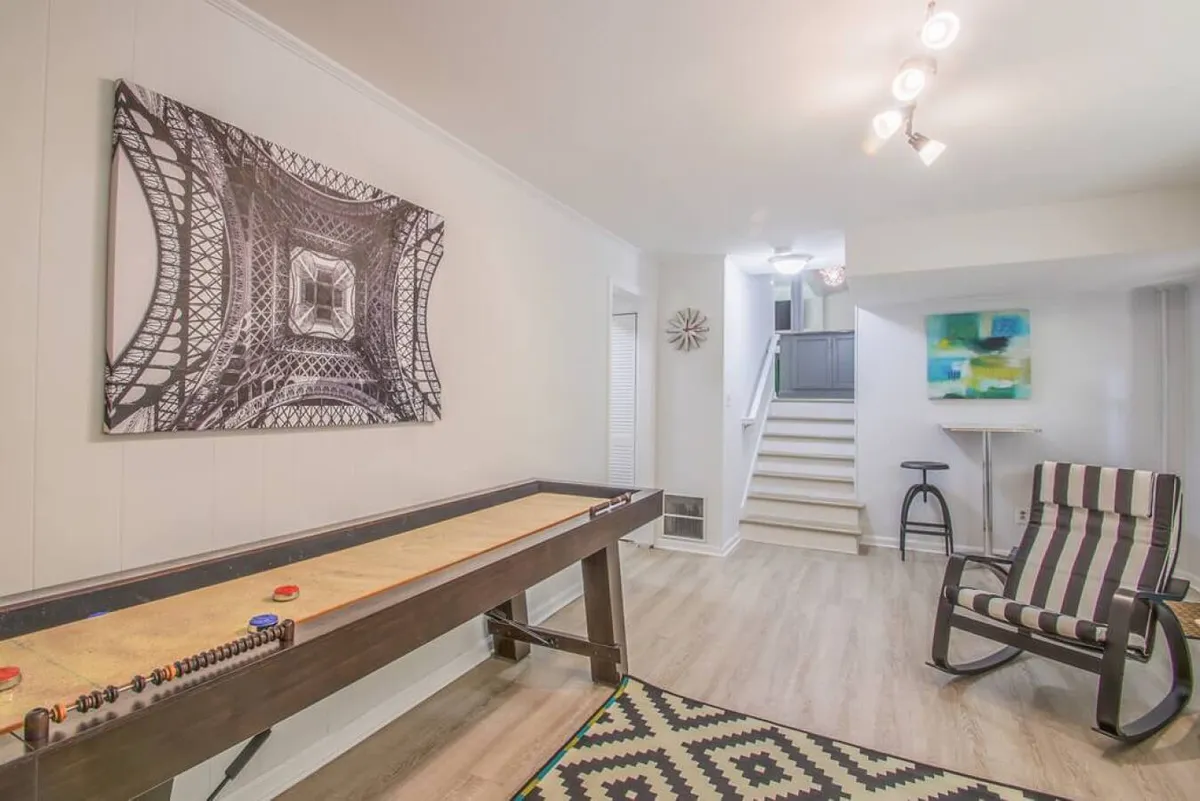 This image features a lively living room with a shuffleboard table as the focal point, complemented by black and white artwork, a striped chair, and a set of stairs leading to the upper level. The space is filled with activity—people playing shuffleboard, relaxing, socializing, and appreciating art—creating an atmosphere of fun and togetherness.