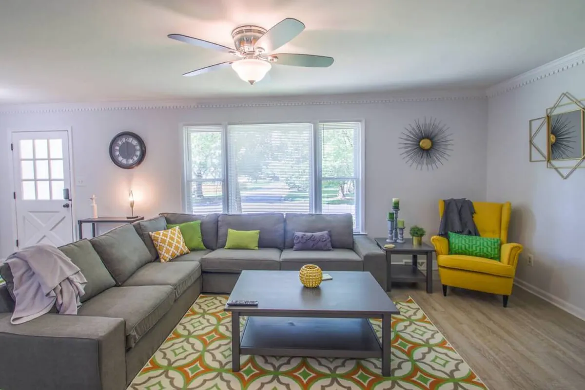 The photo highlights a spacious living room with a large sectional couch, a yellow accent chair, a central coffee table, and expansive windows letting in natural light. The room is set for relaxing with family, hosting guests, watching TV, or reading, and exudes a cozy, vibrant mood.