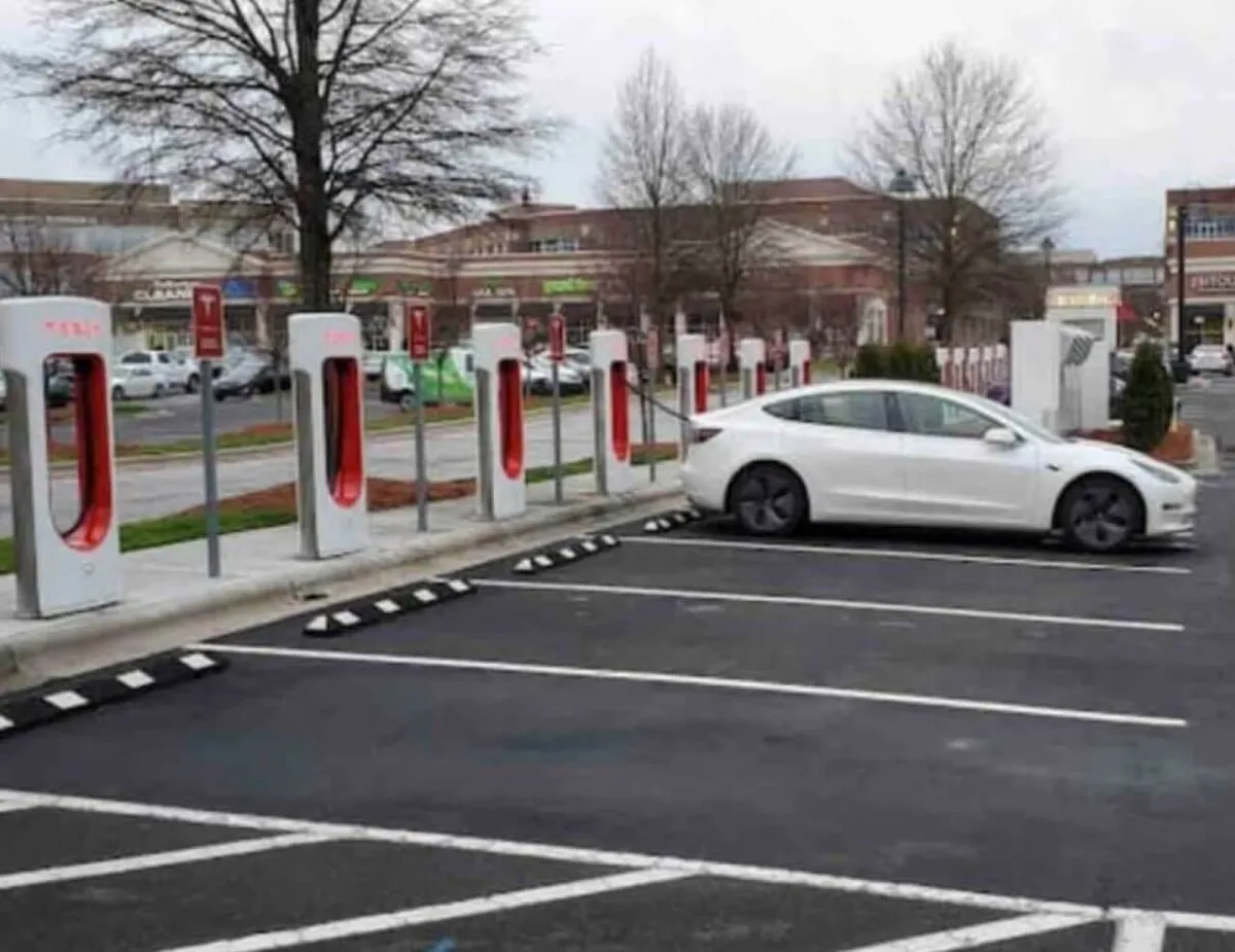 A modern exterior scene featuring Tesla charging stations, a white Tesla vehicle, and a shopping center in the background. The area is bright, clean, and suggests an eco-friendly and business-friendly environment.