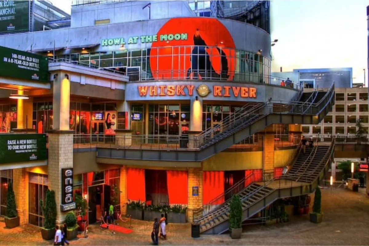 A lively exterior shot capturing the Whisky River sign, a Howl at the Moon advertisement, a staircase leading up, and a bustling outdoor seating area. People are visible socializing and enjoying the energetic nightlife, suggesting the vibrancy of Charlotte’s local dining and entertainment scene.