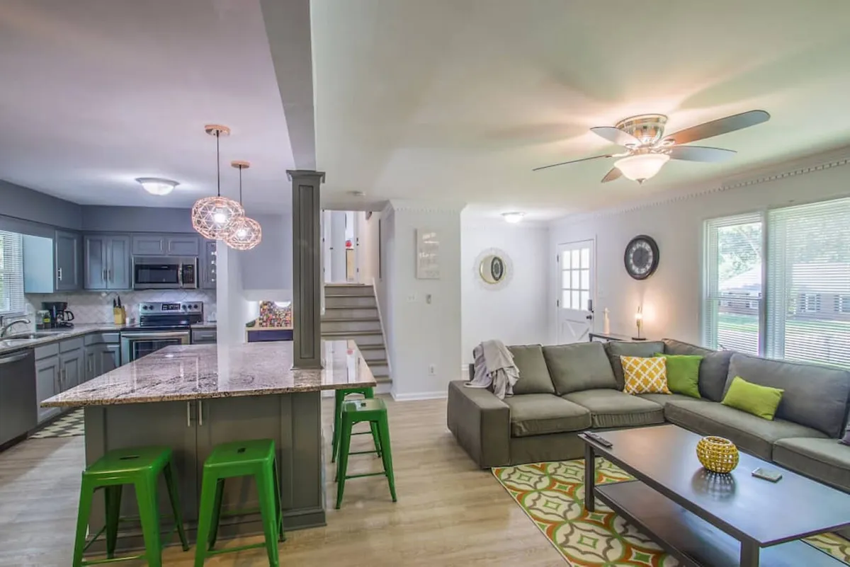 The image showcases a spacious, open-concept living area featuring a stylish kitchen island with contemporary lighting, a plush sofa adorned with decorative pillows, and warm, inviting decor. The scene exudes comfort, perfect for relaxing, enjoying meals, or gathering with family and friends.